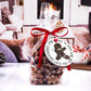 Clear gift bag filled with gingerbread-shaped cookies, tied with a red ribbon. Attached is a round wooden tag with a broken gingerbread man design that reads "Christmas Cost Me an Arm and a Leg." The background shows a cozy living room setting.