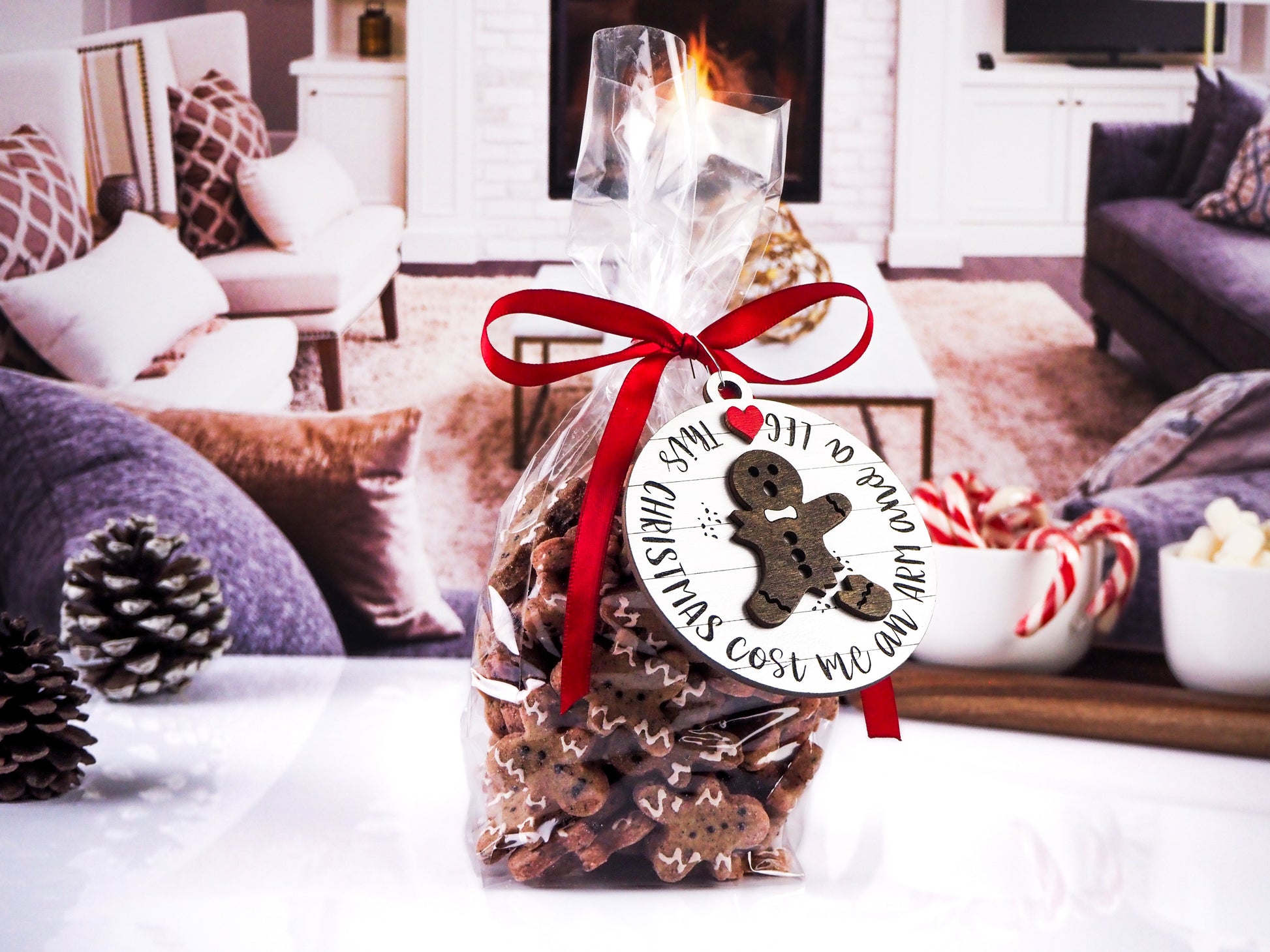 Clear gift bag filled with gingerbread-shaped cookies, tied with a red ribbon. Attached is a round wooden tag with a broken gingerbread man design that reads "Christmas Cost Me an Arm and a Leg." The background shows a cozy living room setting.