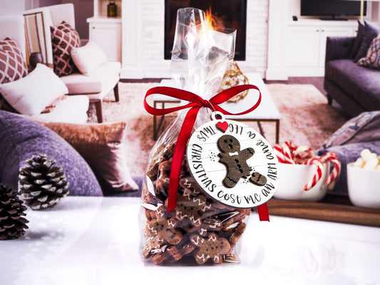 Clear gift bag filled with gingerbread-shaped cookies, tied with a red ribbon. Attached is a round wooden tag with a broken gingerbread man design that reads "Christmas Cost Me an Arm and a Leg." The background shows a cozy living room setting.