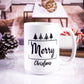 White ceramic mug with "Merry Christmas" printed in black, decorated with various Christmas tree icons above the text. The background shows a cozy living room setup with pinecones and candy canes.