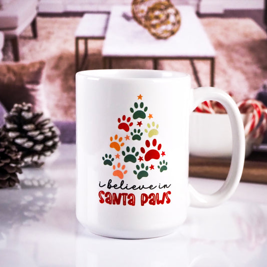 White ceramic mug featuring a Christmas tree shape made up of colorful paw prints, with the text "I believe in Santa Paws" in festive colors. The background includes a cozy living room setup with pinecones and candy canes.