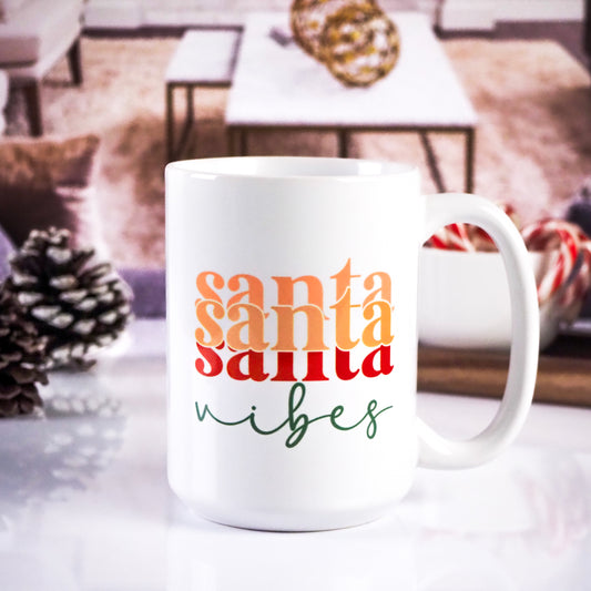 White ceramic mug featuring the phrase "Santa Santa Santa Vibes" in gradient colors of peach, orange, red, and green. The background includes a cozy living room setup with pinecones and candy canes.