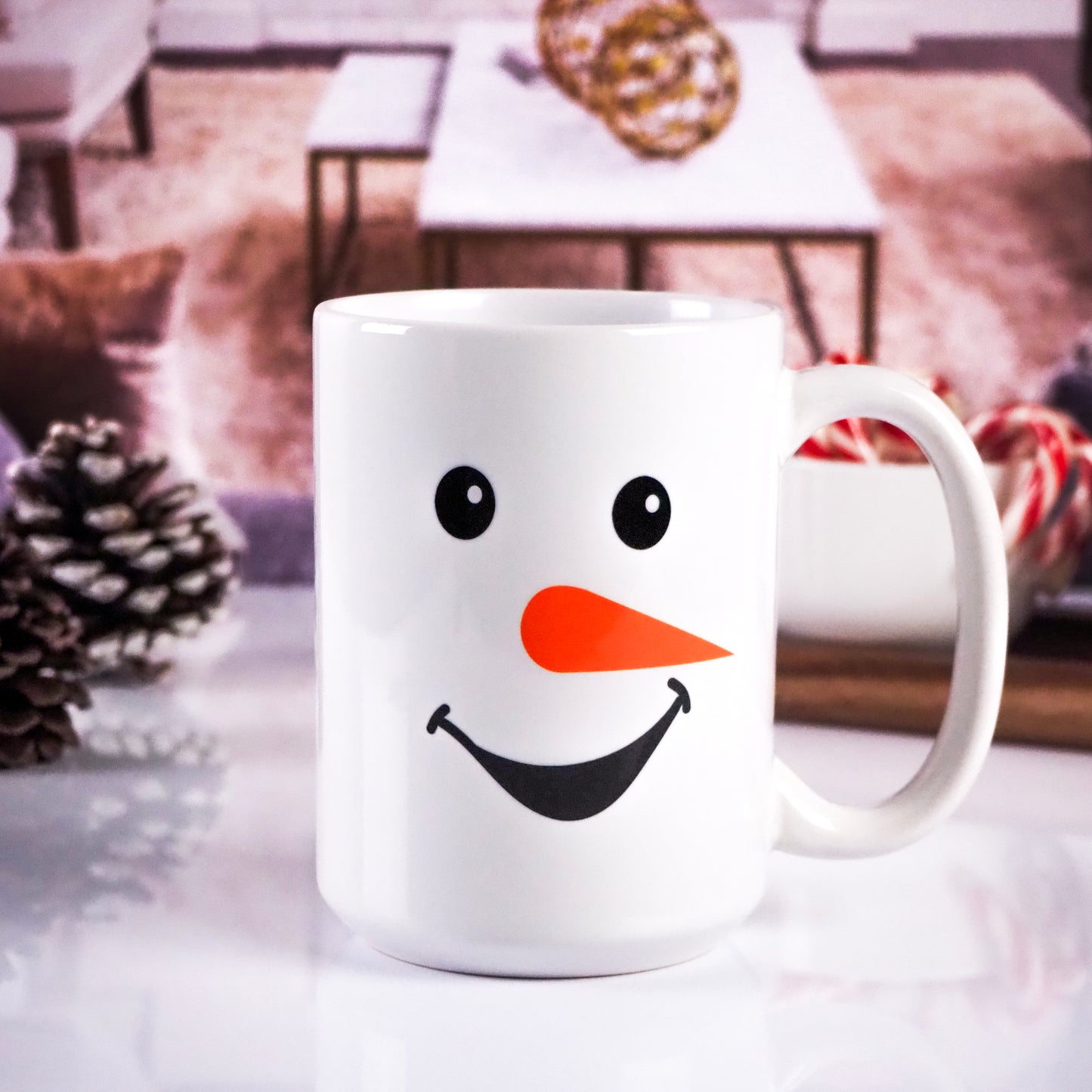 White ceramic mug featuring a smiling snowman design with black eyes, a black mouth, and an orange carrot-shaped nose. Cozy living room setting in the background with pinecones and candy canes.