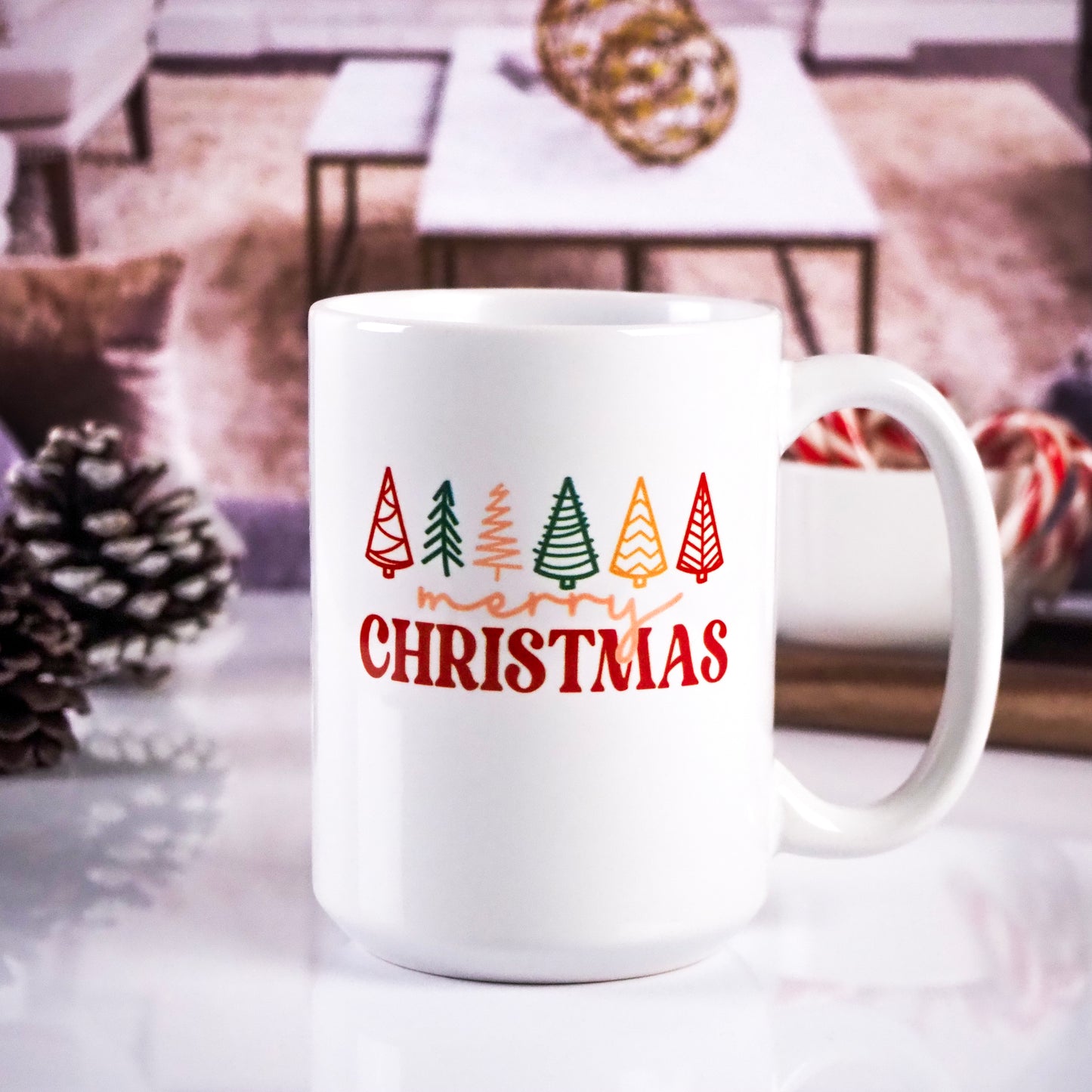 White ceramic mug featuring colorful Christmas trees and the text "Merry Christmas" in red and orange lettering. The background shows a cozy living room setting with pinecones and candy canes.