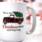 White ceramic mug with the design "This is my Christmas Movie watching mug," featuring a red and black buffalo plaid truck carrying green Christmas trees. The background includes blurred festive holiday décor.