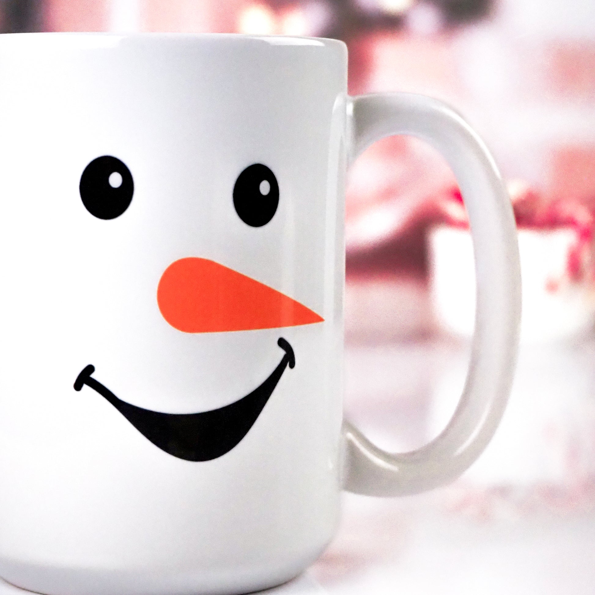 White ceramic mug featuring a smiling snowman design with a carrot nose, black eyes, and a wide smile. The background includes blurred festive holiday décor.