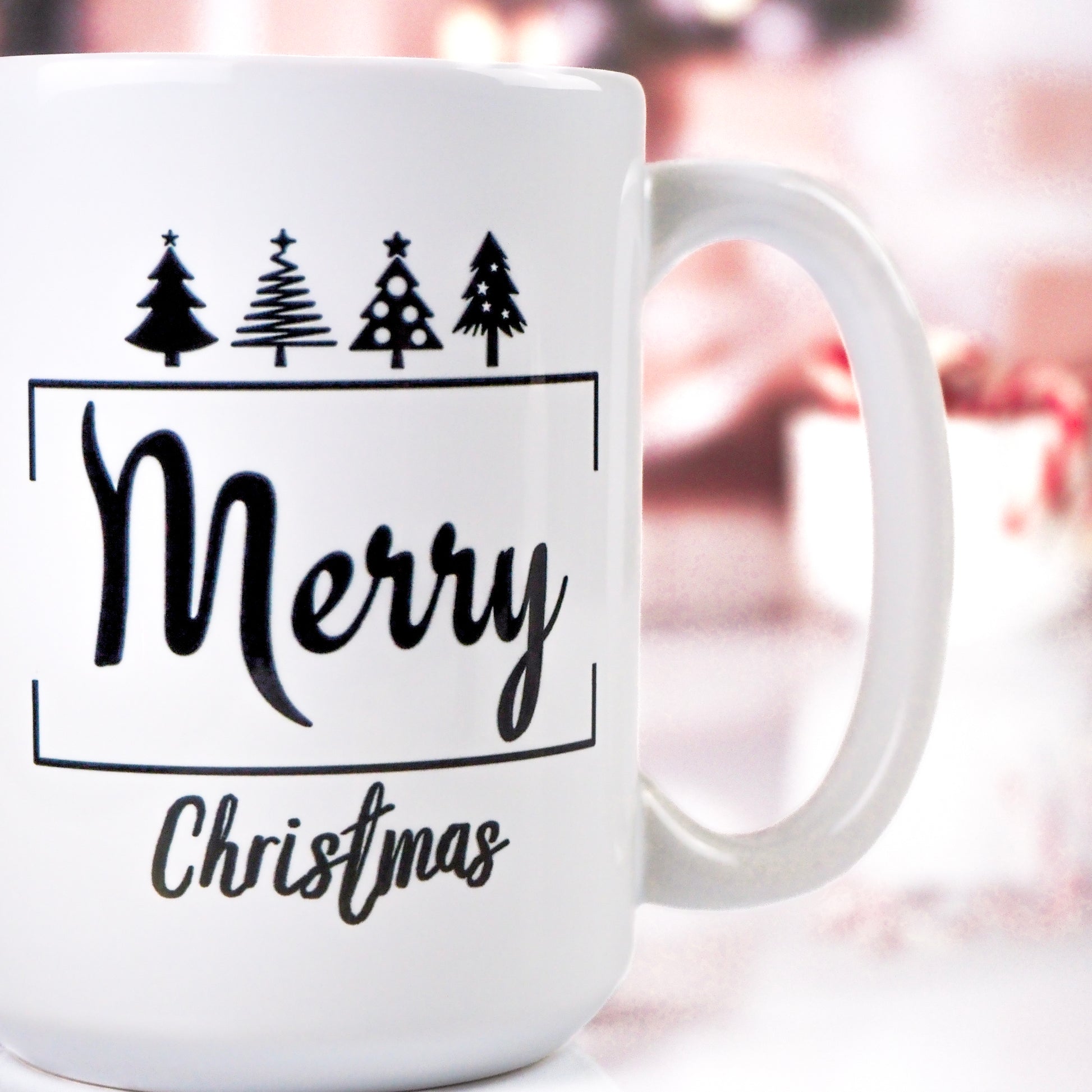 White ceramic mug with "Merry Christmas" written in bold black font, accompanied by small Christmas tree icons at the top. The background features a cozy, blurred holiday scene.