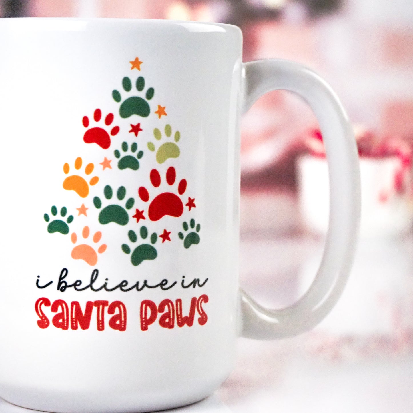 White ceramic mug featuring a Christmas tree made of colorful paw prints, with the phrase "I believe in Santa Paws" in black and red text below. The background includes a cozy, blurred festive setting.