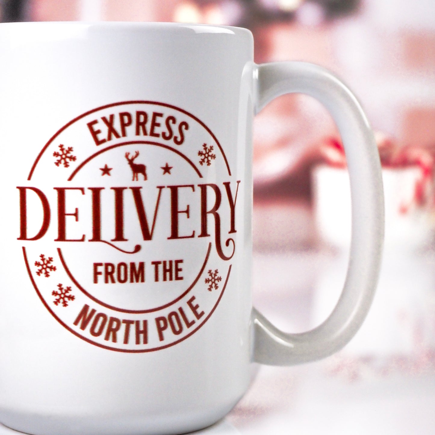 White ceramic mug with a circular red stamp design featuring the text "Express Delivery from the North Pole" and decorative snowflakes, set against a blurred festive background.