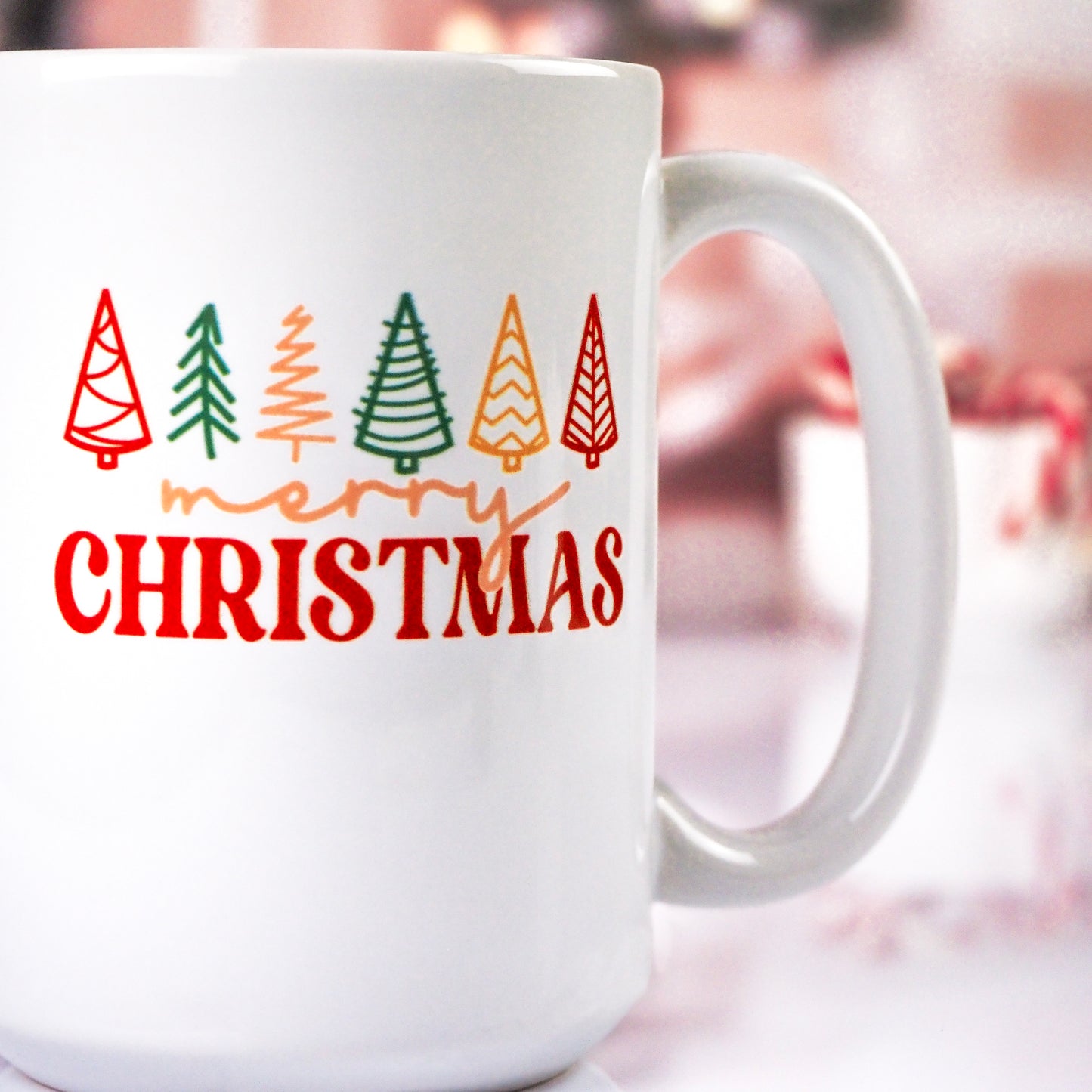 White ceramic mug with a colorful "Merry Christmas" design featuring red, green, yellow, and orange stylized Christmas trees above the text, set against a blurred festive background.