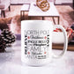 White ceramic mug featuring a festive word collage with phrases like "North Pole," "Jingle Bells," "Cookies," "Nutcracker," "Fireplace," and "Santa," displayed in various fonts and sizes. The mug is set against a cozy, blurred festive background.