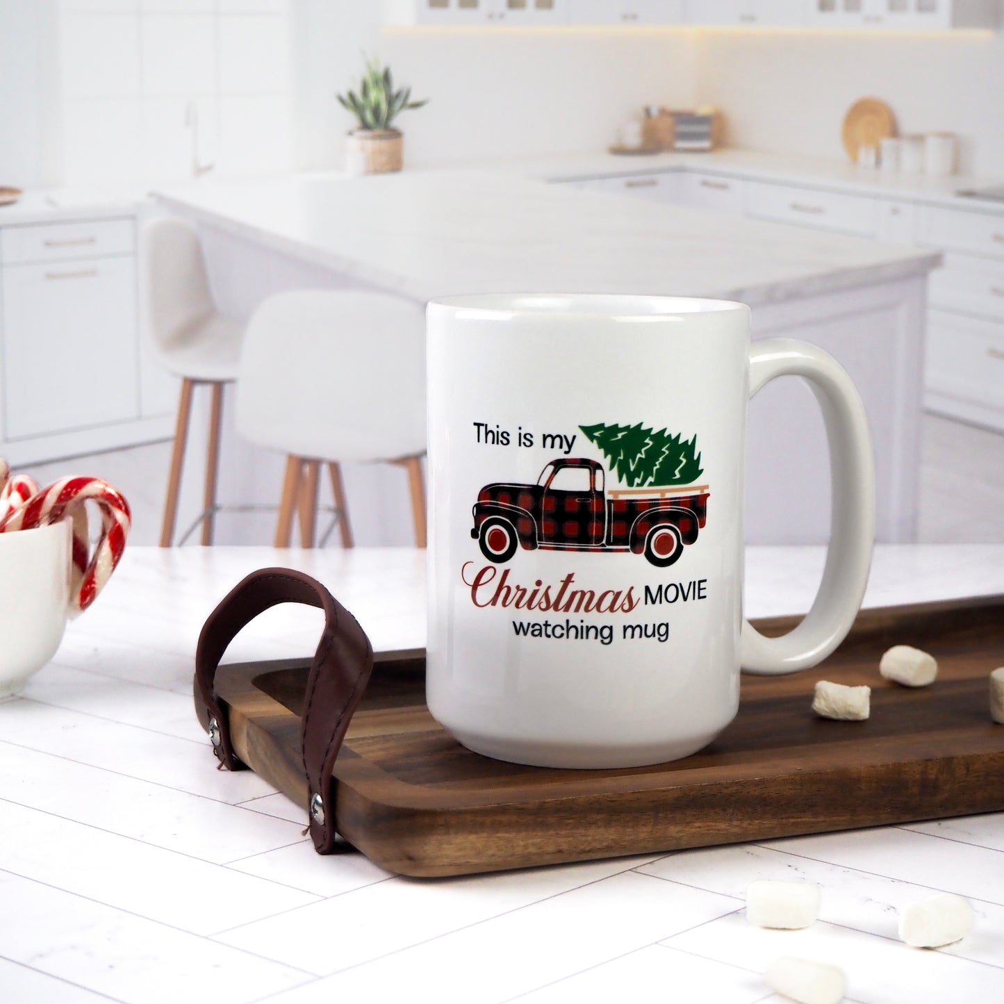 White ceramic mug with a design featuring a red plaid pickup truck carrying a Christmas tree and the phrase "This is my Christmas Movie Watching Mug." The mug is set on a wooden tray, surrounded by marshmallows, candy canes, and a cozy kitchen backdrop.