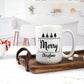 White ceramic mug with the words "Merry Christmas" in black, along with simple black Christmas tree illustrations. The mug is placed on a wooden tray with marshmallows and a cozy kitchen setting, featuring a candy cane in the background.