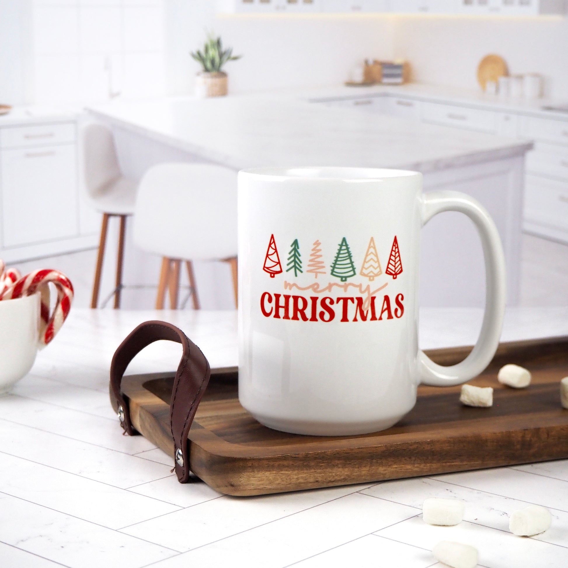 White ceramic mug featuring "Merry Christmas" in red with colorful Christmas tree illustrations above. The mug sits on a wooden tray, surrounded by marshmallows, candy canes, and a bright kitchen backdrop.