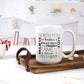 White ceramic mug featuring a word collage design with festive words like "North Pole," "Christmas," "Family," "Jingle Bells," and more in black and red text. The mug sits on a wooden tray, surrounded by marshmallows and a bright kitchen setting.