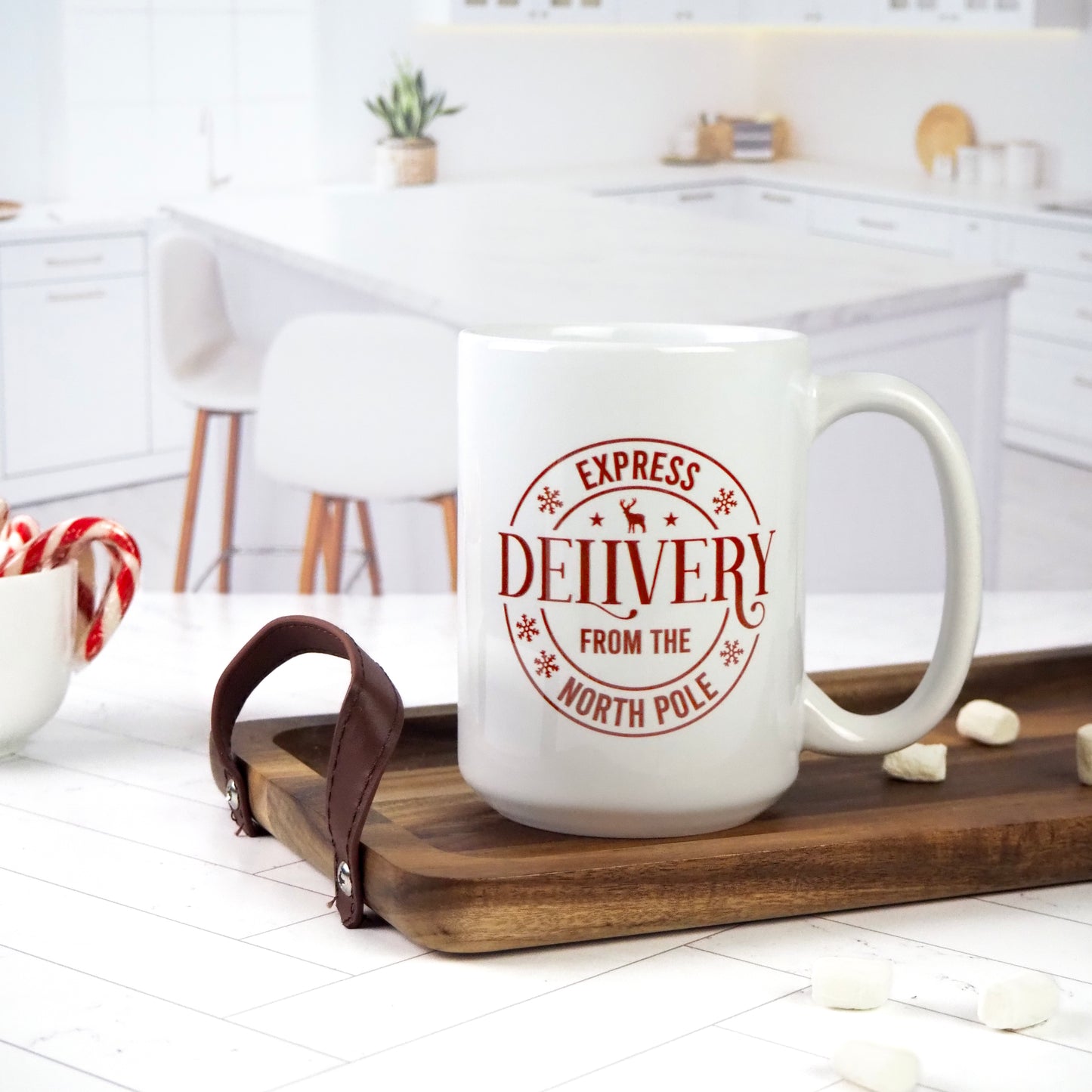 White ceramic mug featuring the text "Express Delivery from the North Pole" in a red, circular stamp-like design. The mug sits on a wooden tray in a bright kitchen setting, surrounded by marshmallows and candy canes.