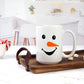 White ceramic mug featuring a cute snowman face design with a bright orange carrot nose and a smiling mouth. The mug sits on a wooden tray in a bright kitchen setting, with marshmallows and candy canes nearby.