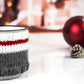 White with black speckles campfire mug wrapped in a knitted cozy with white, red, and gray stripes. The mug is placed on a white surface with red Christmas ornaments and a pine cone in the background, creating a warm, festive ambiance.