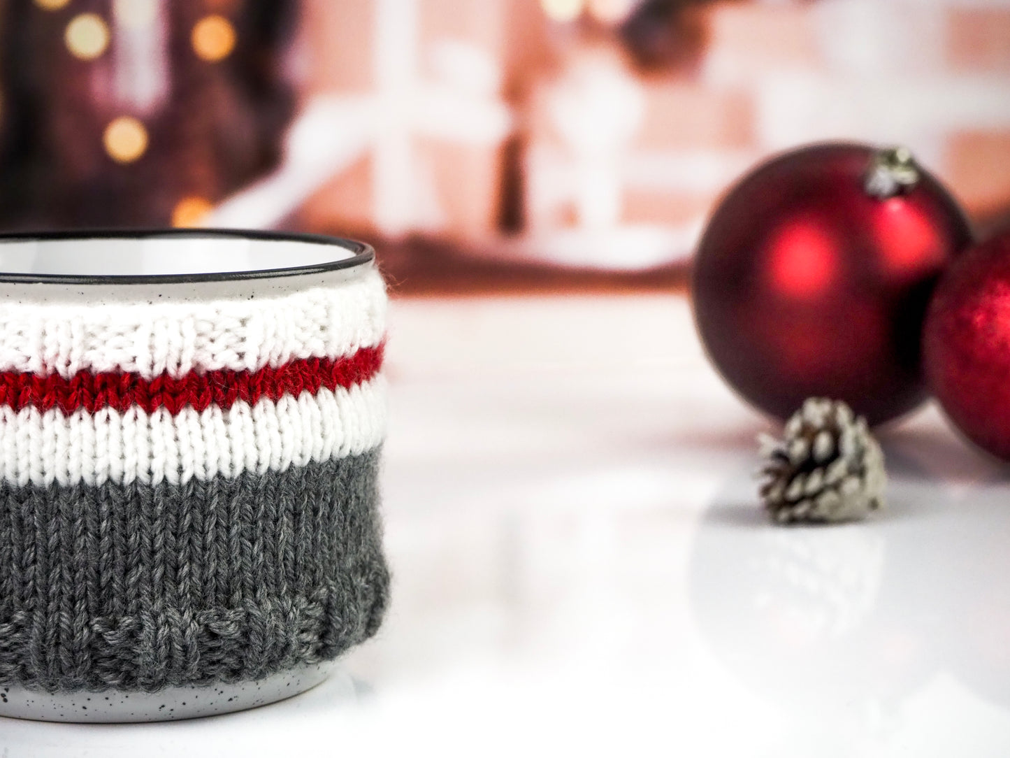 White with black speckles campfire mug wrapped in a knitted cozy with white, red, and gray stripes. The mug is placed on a white surface with red Christmas ornaments and a pine cone in the background, creating a warm, festive ambiance.