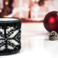 White with black speckles  campfire mug wrapped in a black and white knitted cozy with a snowflake pattern, set against a festive background with red ornaments and a pine cone.