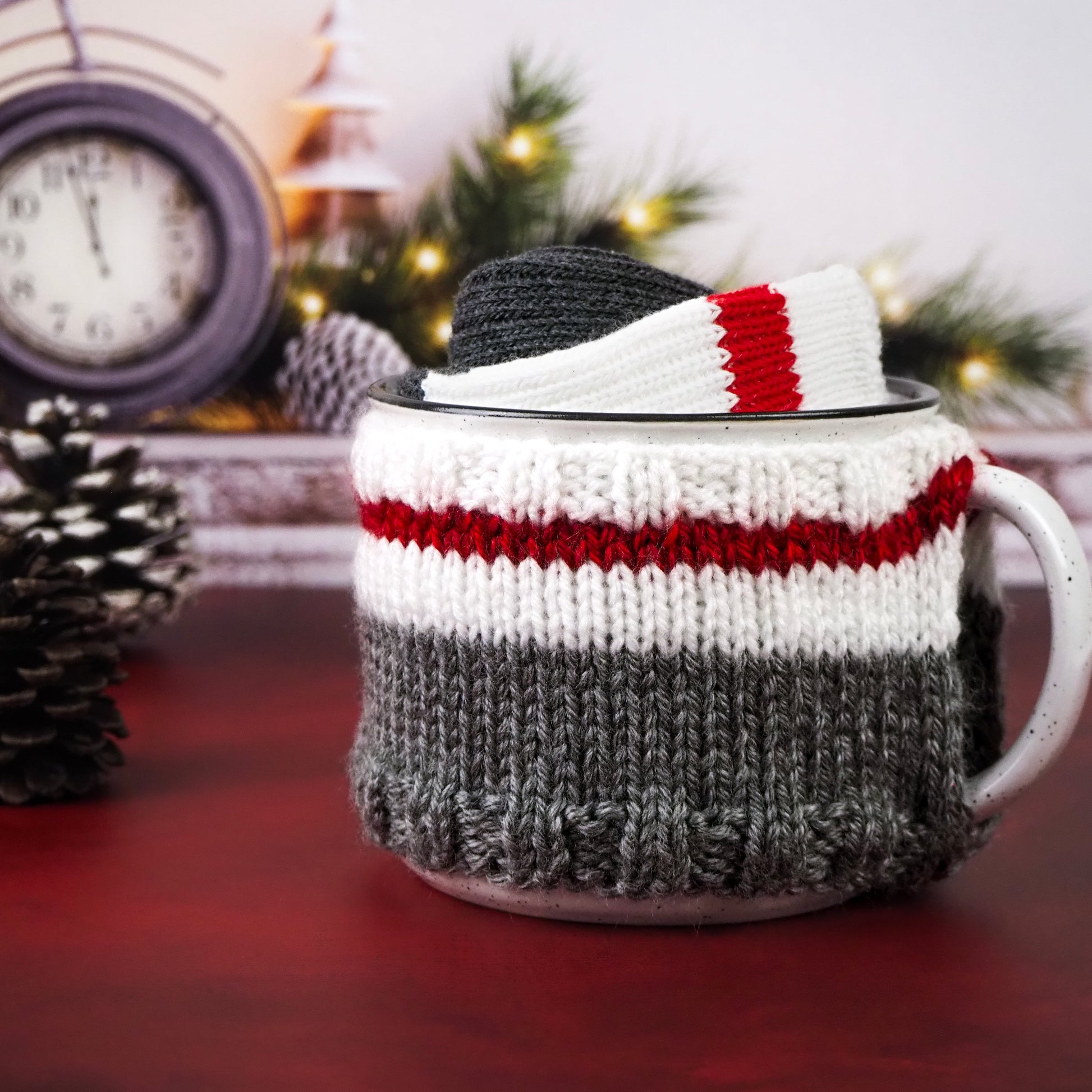 White with black speckles campfire mug wrapped in a knitted cozy with white, red, and gray stripes and matching socks inside the mug. The mug is set against a red surface, with additional cozy material inside the mug. A pine cone, a clock, and a festive background with lit Christmas trees complete the scene.