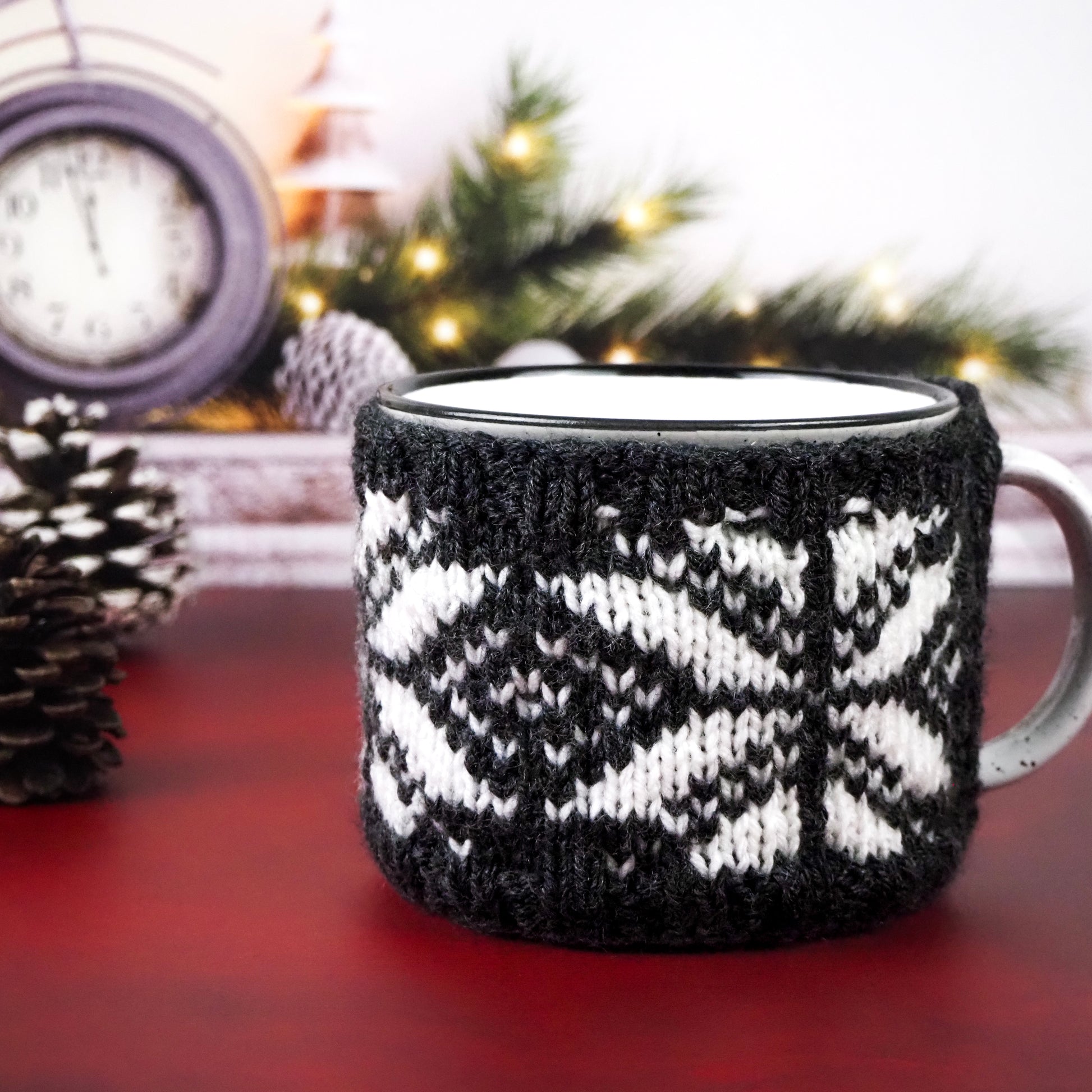 White with black speckles  campfire mug with a black and white snowflake-patterned knitted cozy, set on a red surface with festive pine branches and a clock in the background.