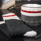 White with black speckles campfire mug wrapped in a knitted cozy with white, red, and gray stripes, placed alongside a pair of matching knit socks. The set is displayed on a glossy black surface with a blurred book background.