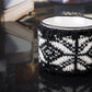 Close-up of a white with black speckles campfire mug wrapped in a black and white snowflake-patterned knitted cozy, with a dark reflective surface and blurred background details.