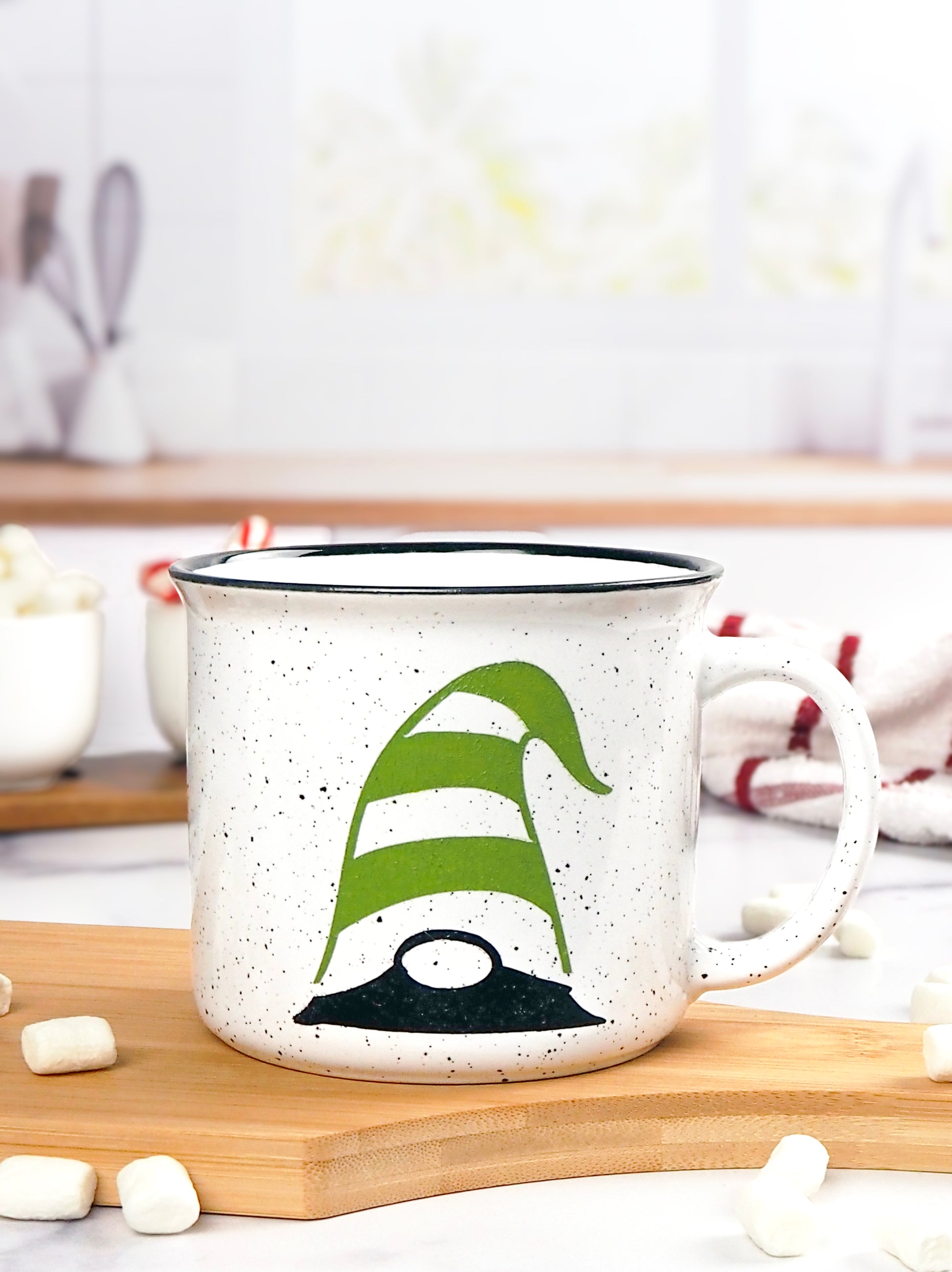 White speckled ceramic campfire mug with a black rim. The mug features a whimsical gnome design, with the gnome wearing a green and white striped hat that covers its eyes, with only the black nose and part of the beard visible.