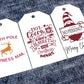 Three white rectangular Christmas gift tags. The first tag features the words North Pole Express Mail in red, with a gold reindeer in the center. The second tag reads Hot Cocoa, Cozy Blankets, and Christmas Movies in red, surrounded by festive icons like a mug, snowflakes, and hearts. The third tag shows a gnome holding a sign that says No Peeking!, with Merry Christmas written in black script at the bottom.