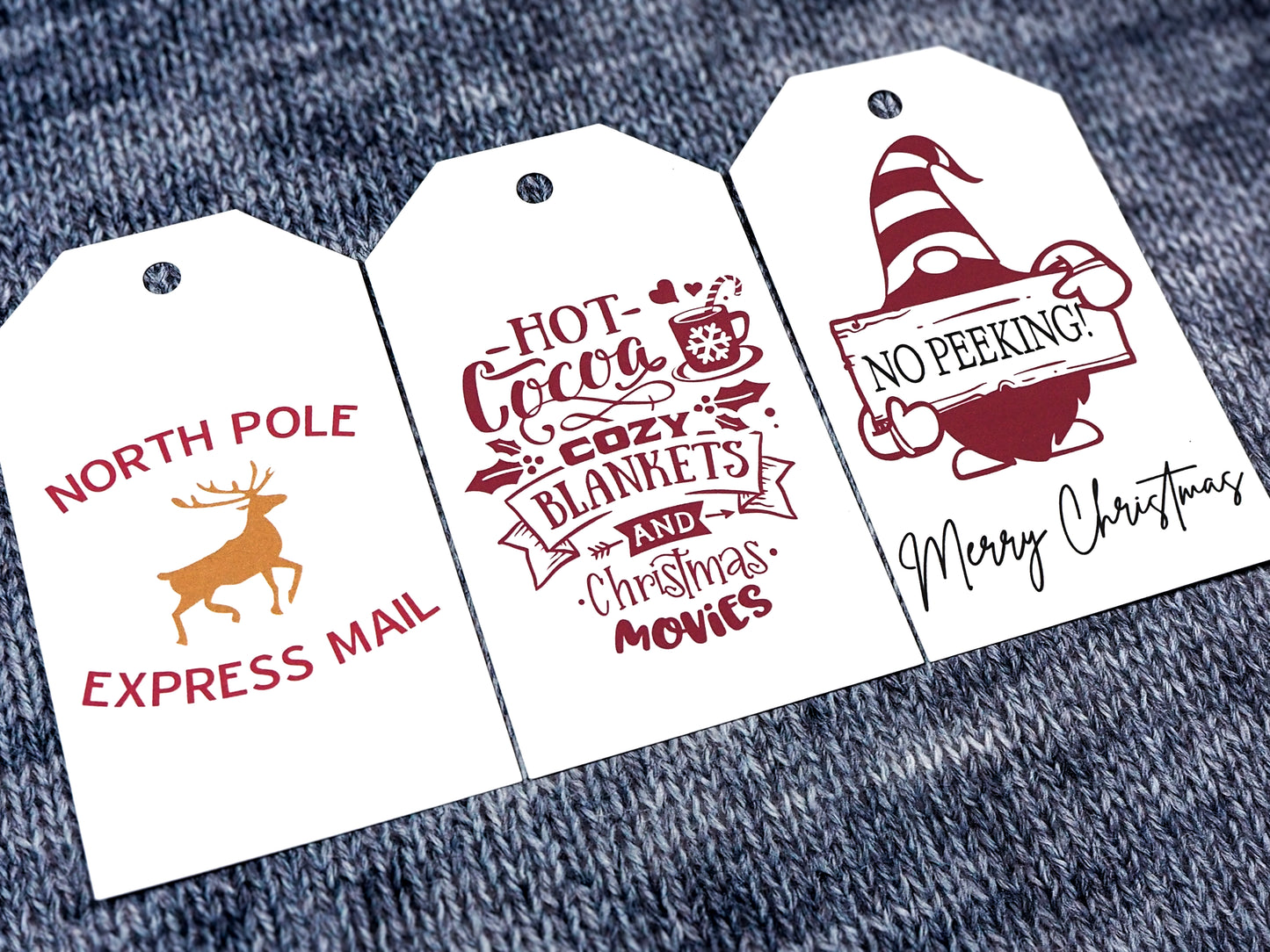 Three white rectangular Christmas gift tags. The first tag features the words North Pole Express Mail in red, with a gold reindeer in the center. The second tag reads Hot Cocoa, Cozy Blankets, and Christmas Movies in red, surrounded by festive icons like a mug, snowflakes, and hearts. The third tag shows a gnome holding a sign that says No Peeking!, with Merry Christmas written in black script at the bottom.