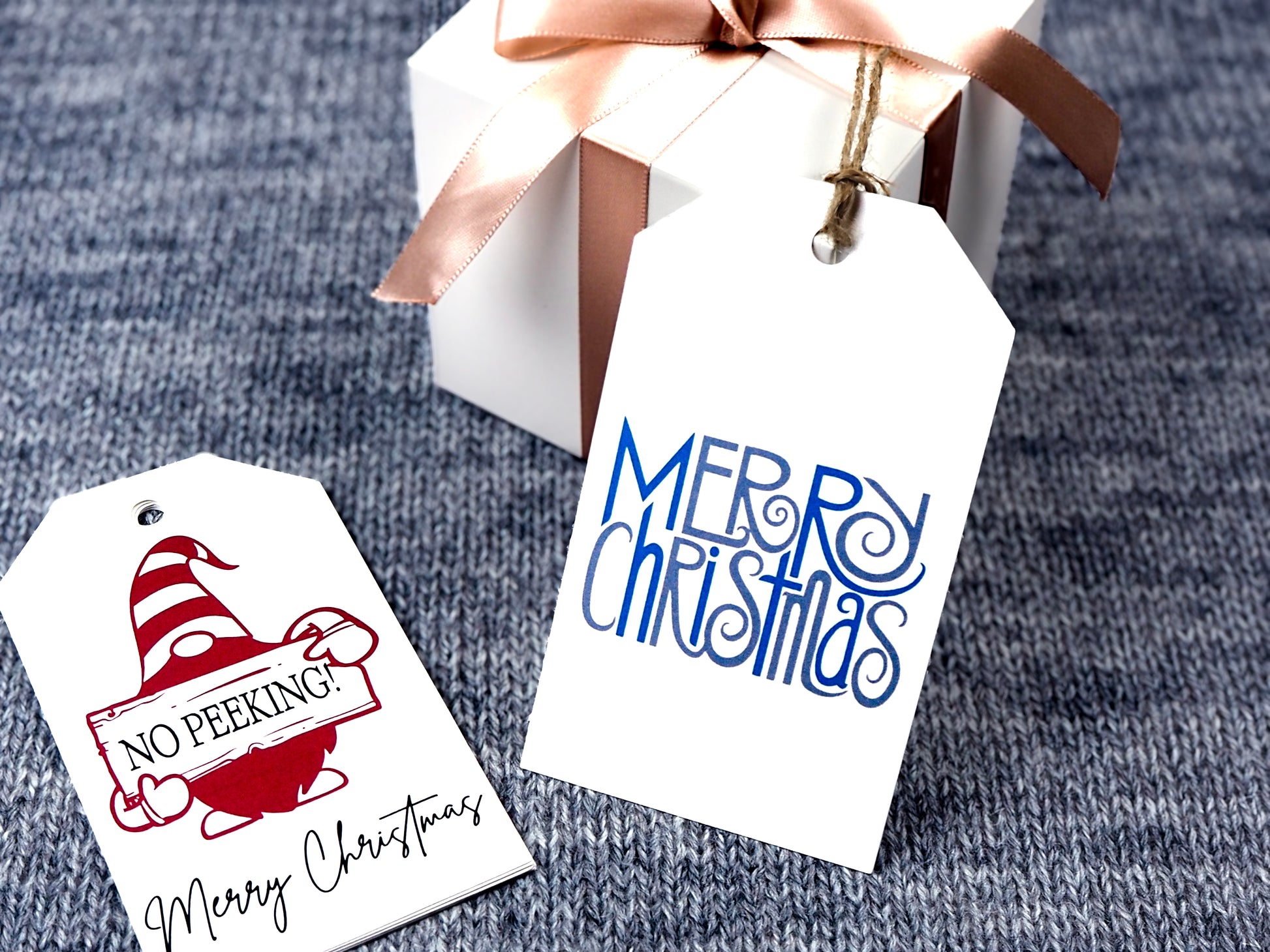 Two white rectangular Christmas gift tags. The first tag features a red gnome holding a sign that says No Peaking! with Merry Christmas written in black script at the bottom. The second tag has the phrase Merry Christmas written in a whimsical blue and gray font.