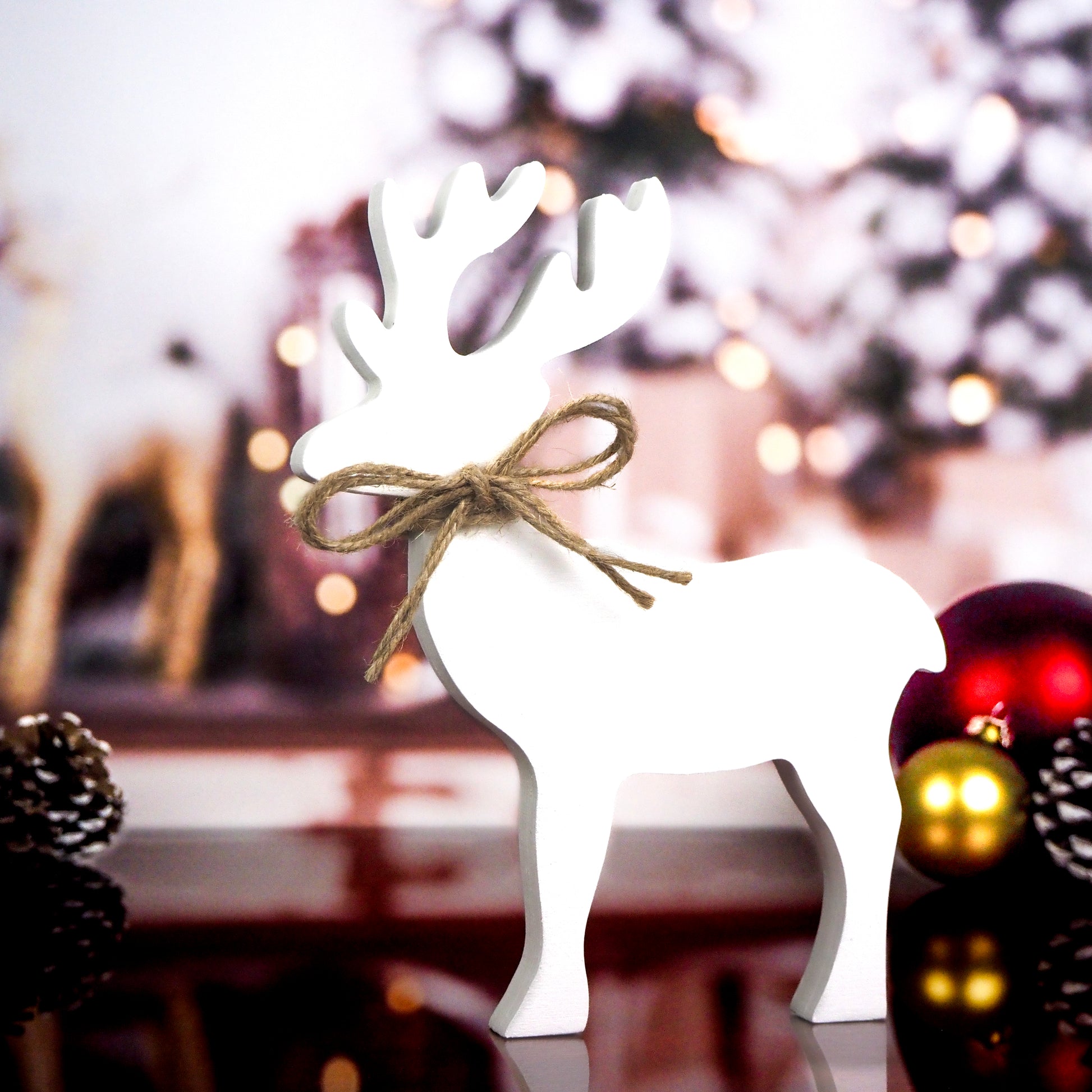 White wooden reindeer with a twine bow around its neck, standing against a festive holiday background with blurred lights and ornaments.