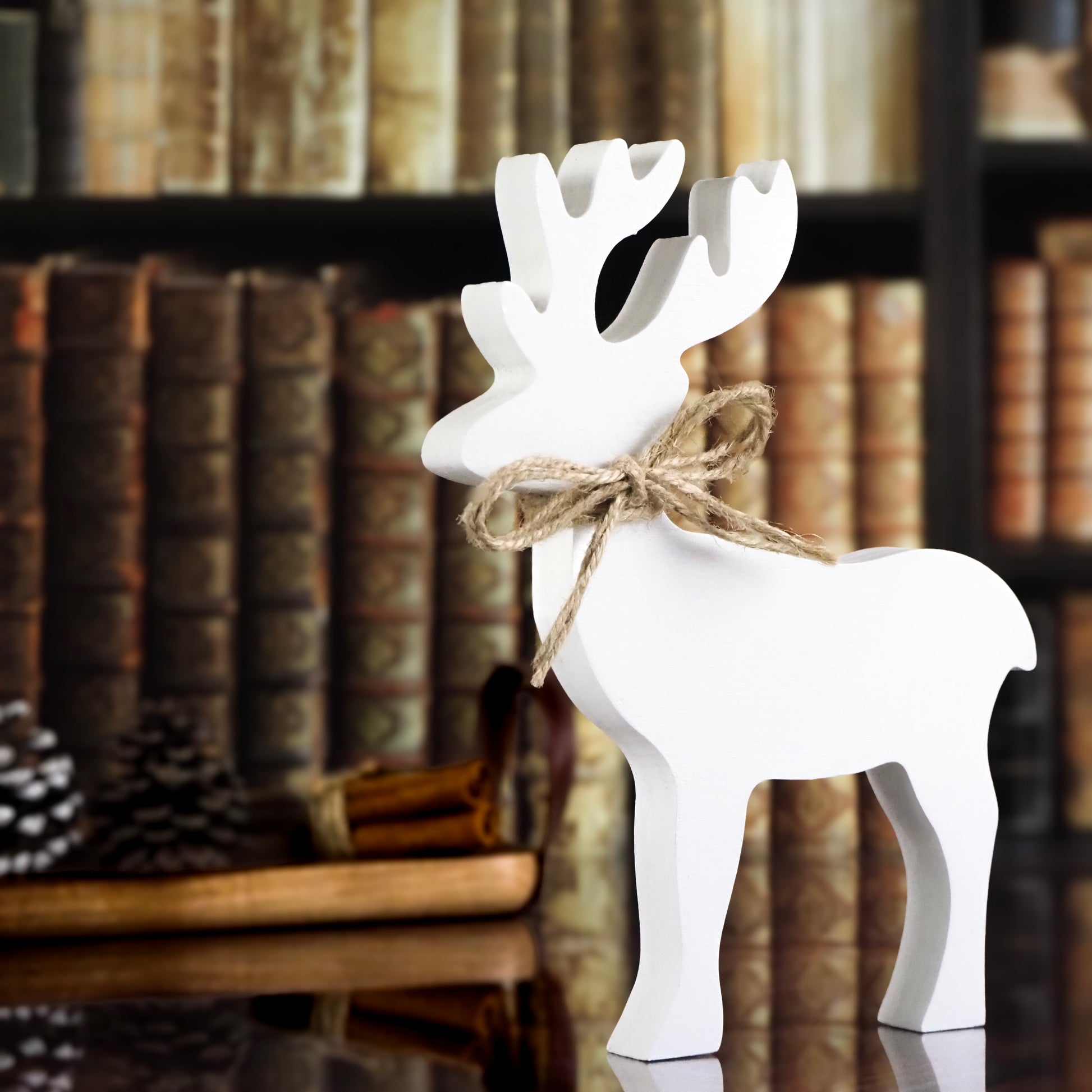 White wooden reindeer with a twine bow around its neck, displayed against a backdrop of vintage books and cozy pinecones.