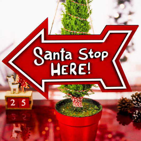 Red arrow-shaped sign with 'Santa Stop Here!' text, hanging in front of a festive potted tree and holiday décor.