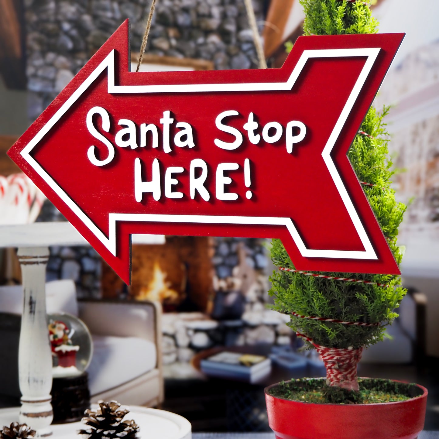 Red arrow-shaped 'Santa Stop Here!' sign hanging in front of a festive potted tree with a cozy holiday-themed interior background.