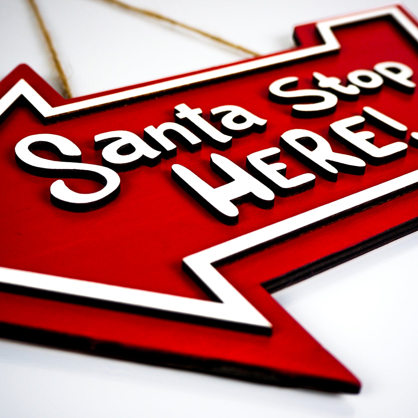 Close-up angled view of a red 'Santa Stop Here!' arrow-shaped sign with bold white lettering and a twine hanging loop.