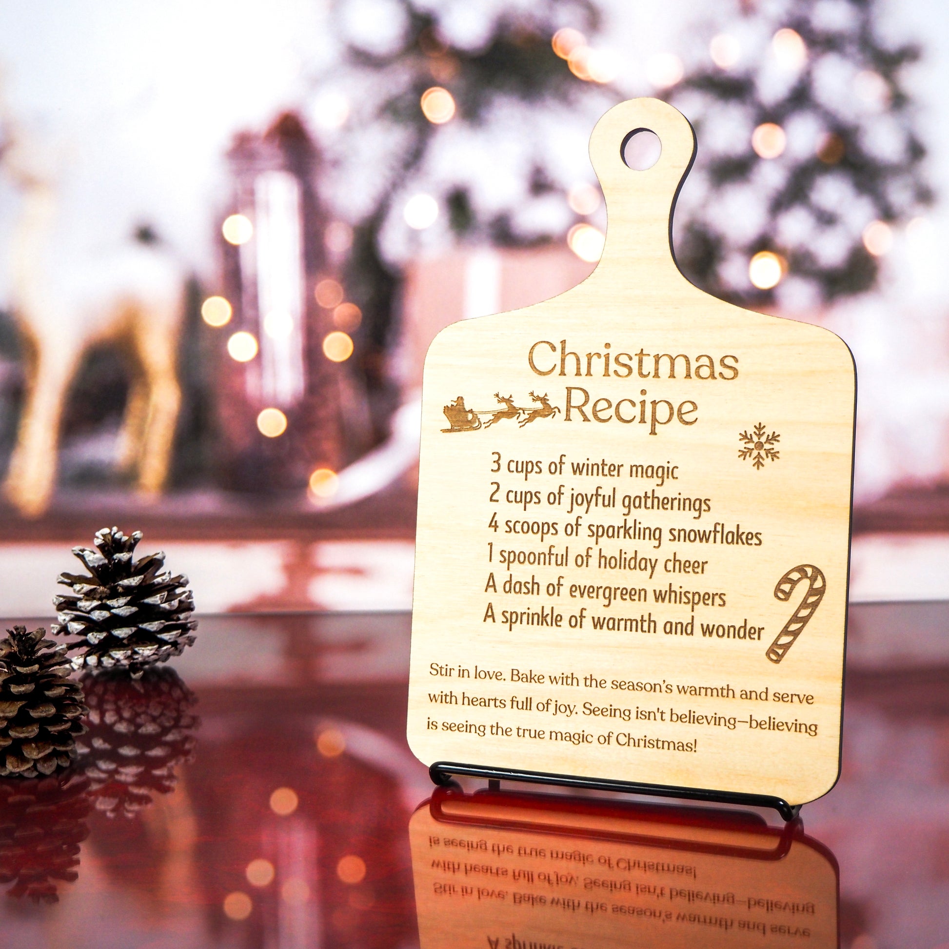 Wooden cutting board-style decoration engraved with a festive Christmas recipe, displayed upright on a stand with a blurred holiday-themed background featuring pinecones and soft twinkle lights.