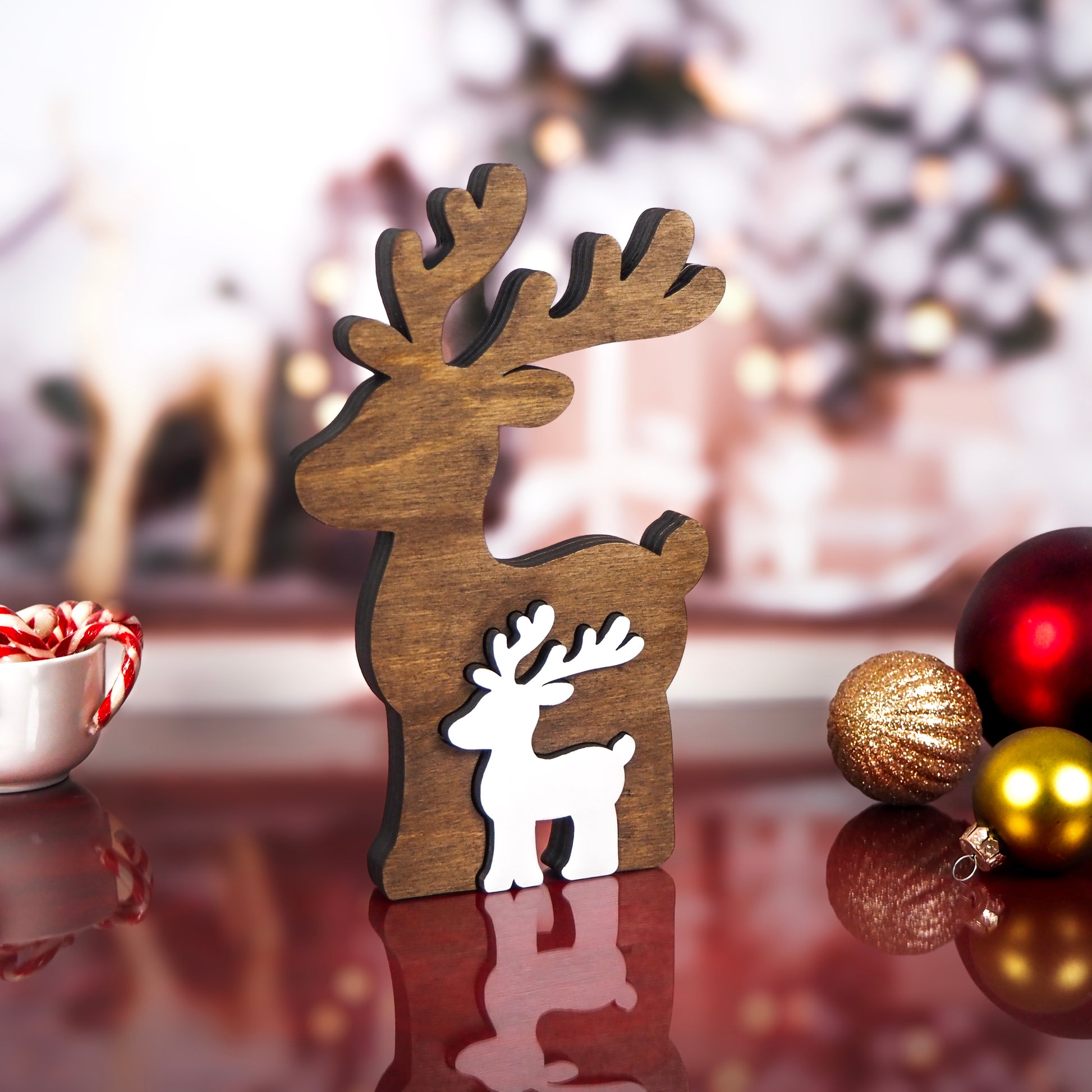 Wooden reindeer decoration with a rustic brown finish, featuring a smaller white reindeer nested inside, displayed on a reflective red table with Christmas ornaments and candy canes in the background.