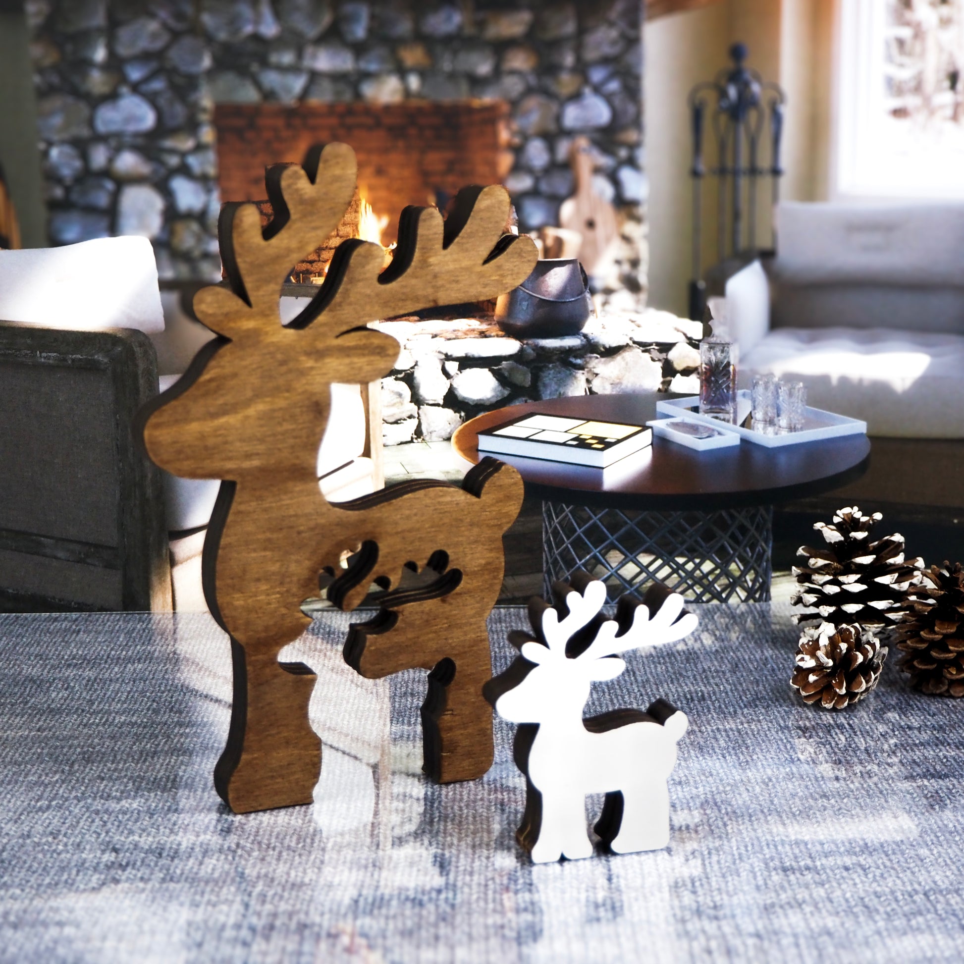 Set of two wooden reindeer decorations, one in rustic brown and one in white, placed on a cozy living room backdrop with a stone fireplace, pinecones, and a coffee table.