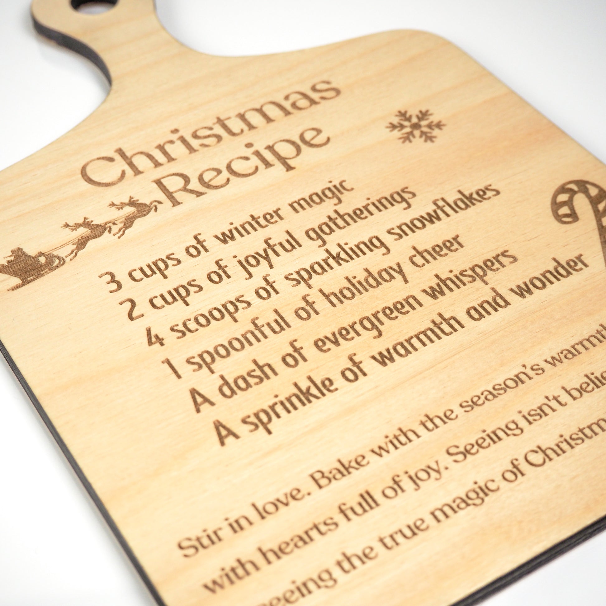 Close-up view of a wooden cutting board-style decoration engraved with a festive Christmas recipe, featuring detailed text and holiday-themed illustrations such as a sleigh, snowflake, and candy cane.