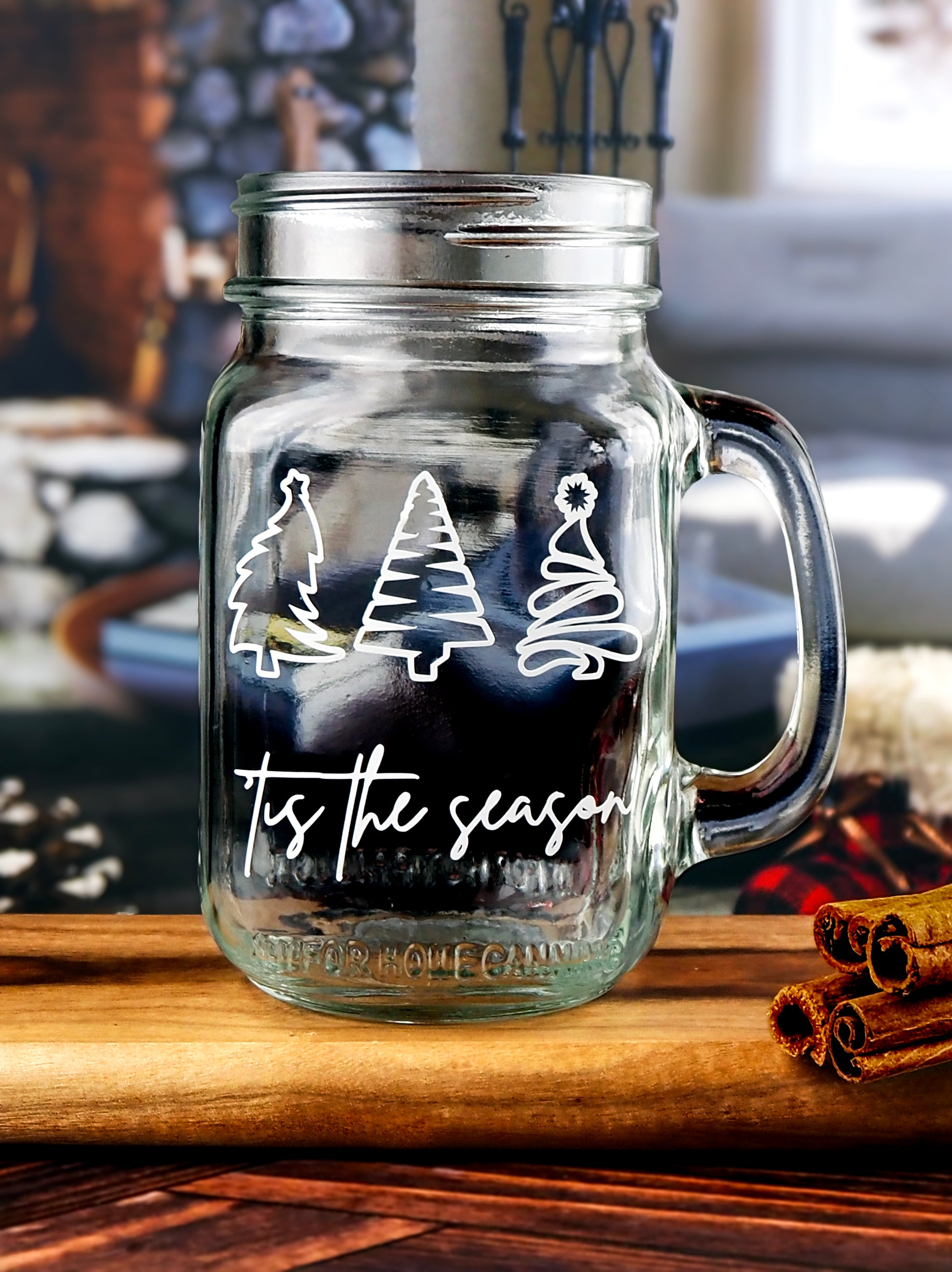 Clear glass mason jar-style mug with a handle. The mug features three white, stylized Christmas trees in a row at the top. Below the trees, the text 'Tis the season is written in a white, flowing script.