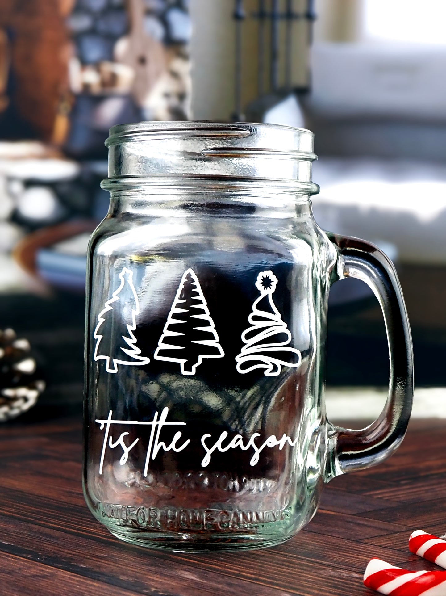 Clear glass mason jar-style mug with a handle. The mug features three white, stylized Christmas trees in a row at the top. Below the trees, the text 'Tis the season is written in a white, flowing script.