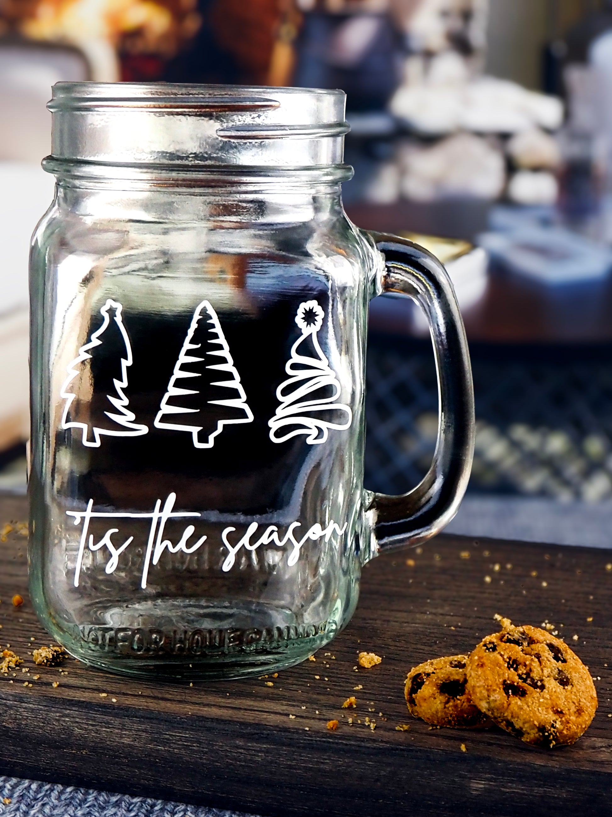 Clear glass mason jar-style mug with a handle. The mug features three white, stylized Christmas trees in a row at the top. Below the trees, the text 'Tis the season is written in a white, flowing script.