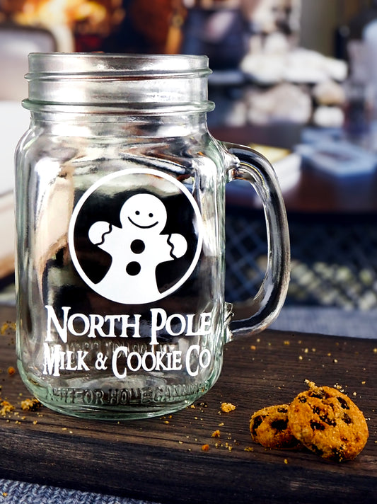 Clear glass mason jar-style mug with a handle. The mug features a white graphic of a smiling gingerbread man inside a circle, along with the text North Pole Milk & Cookie Co in a vintage-style font.