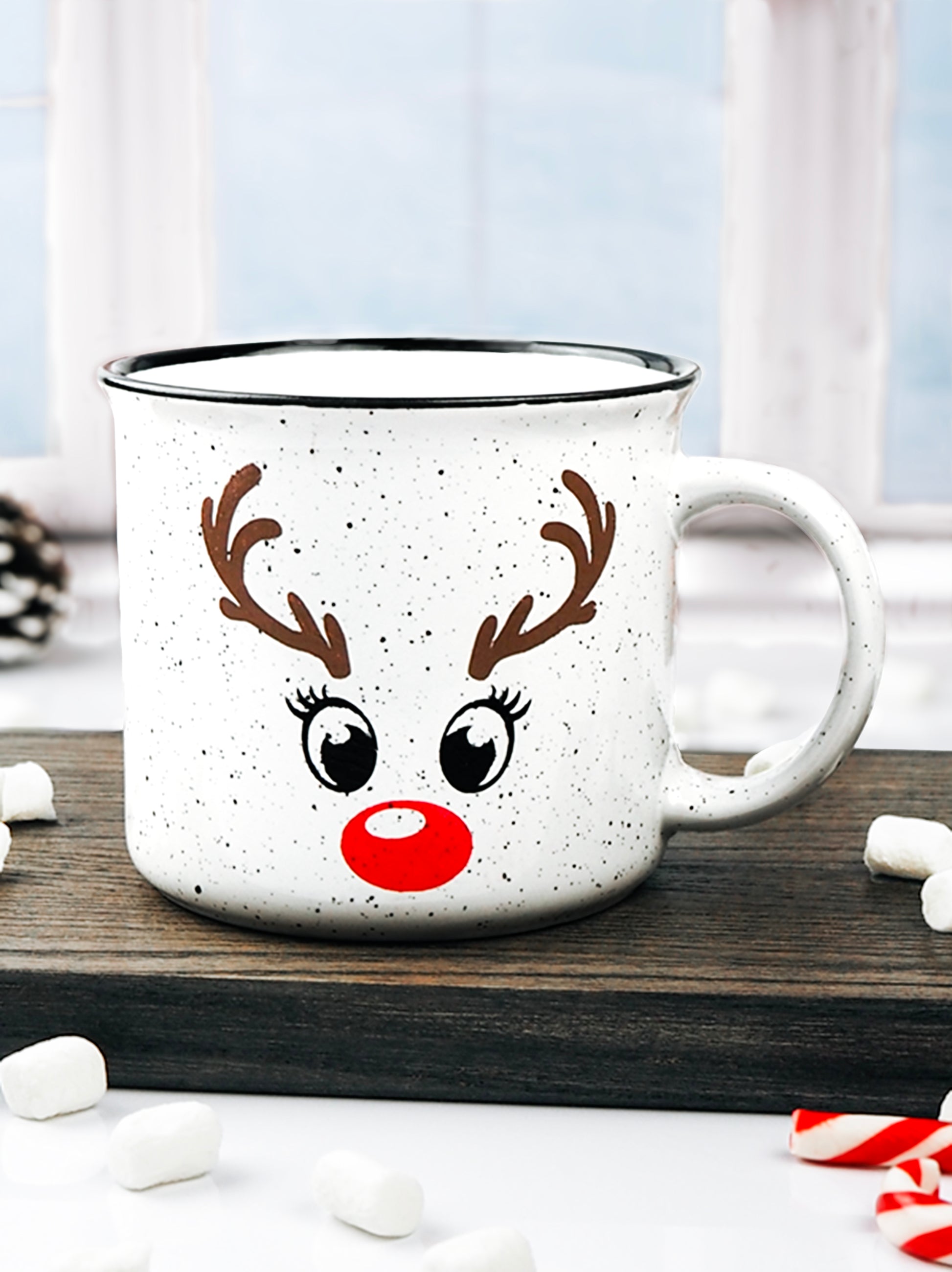 White speckled ceramic campfire mug with a black rim, featuring a reindeer face design. The face includes large black eyes, brown antlers, and a bright red nose, resembling Rudolph the Red-Nosed Reindeer.