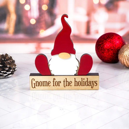 A festive gnome holiday tabletop decoration with a red hat, white beard, and wooden base featuring the text "Gnome for the holidays." The gnome is placed on a white surface with Christmas ornaments and a pinecone in the background.