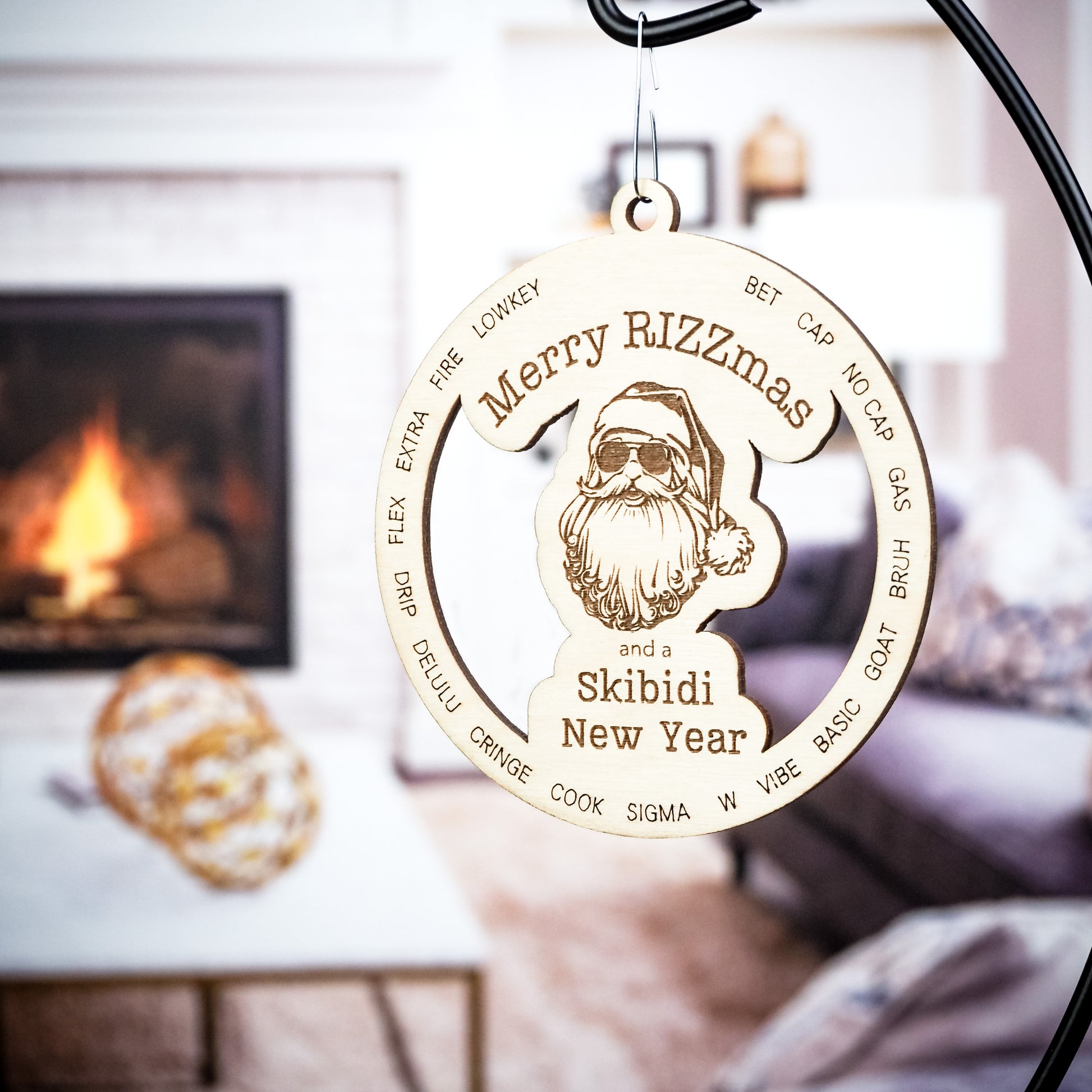 Wooden Christmas ornament engraved with Merry Rizzmas and a Skibidi New Year featuring a Santa wearing sunglasses. Trendy slang words are etched around Santa in a circular design. The ornament is displayed on a black metal stand with a cozy living room in the background, including a lit fireplace and blurred furniture.