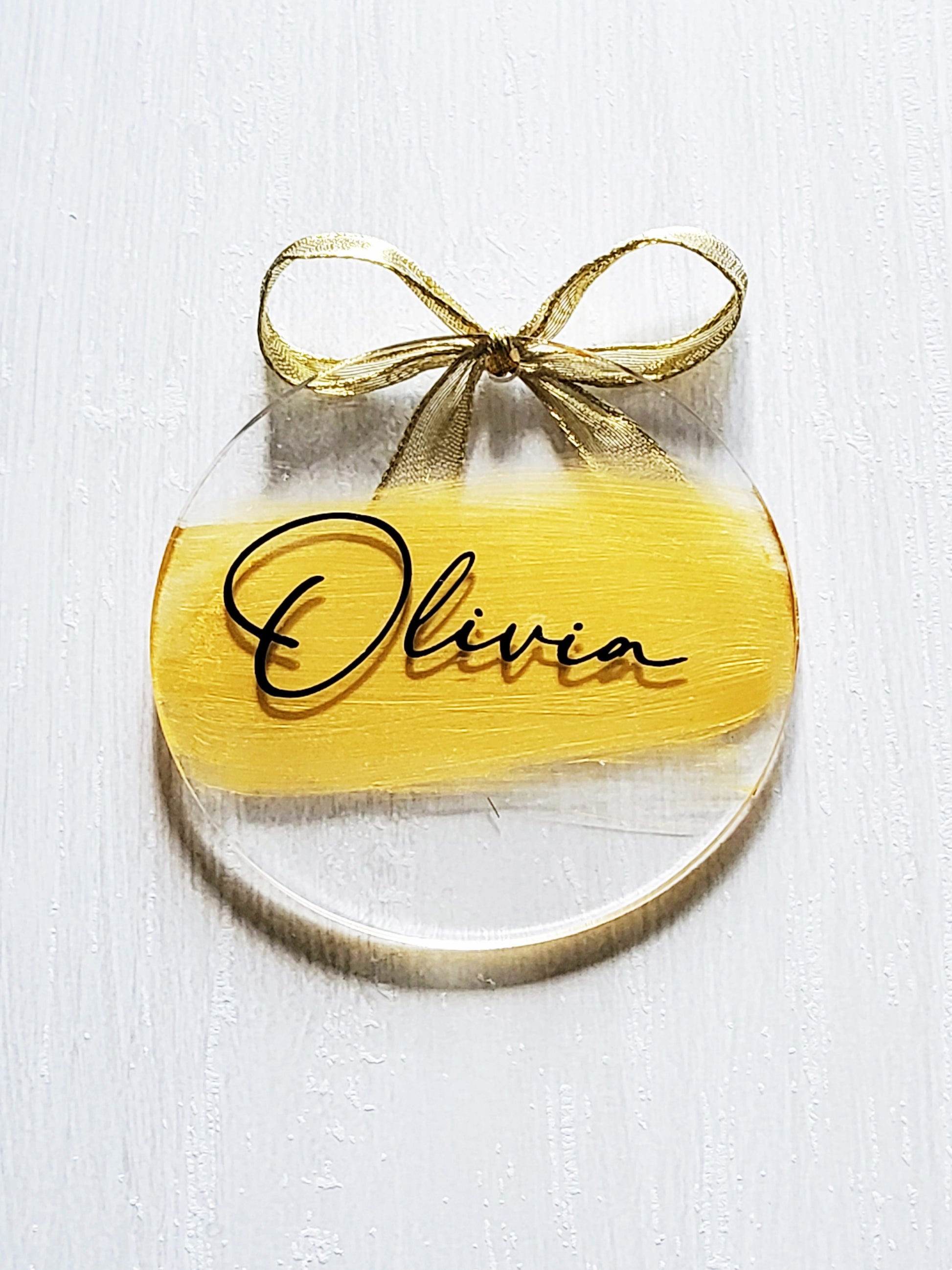 3-inch acrylic discs with gold hand-painted on the back, a personalized name written on the front in black and a gold ribbon tied in a bow. Handmade by Snuggle Up Co.