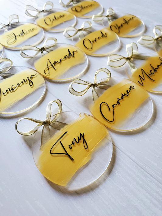 3-inch acrylic discs with gold hand-painted on the back, a personalized name written on the front in black and a gold ribbon tied in a bow. Handmade by Snuggle Up Co.